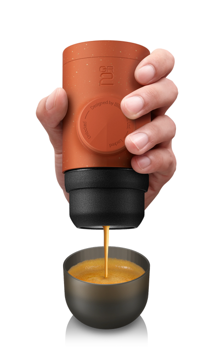 Hand holding a terracotta-colored portable coffee maker dispensing coffee into a small cup.